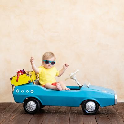 Toddler wearing sunglasses sitting in a toy car with a suitcase on the back