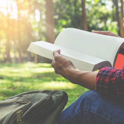 Person reading a book near a forest with a backpack nearby